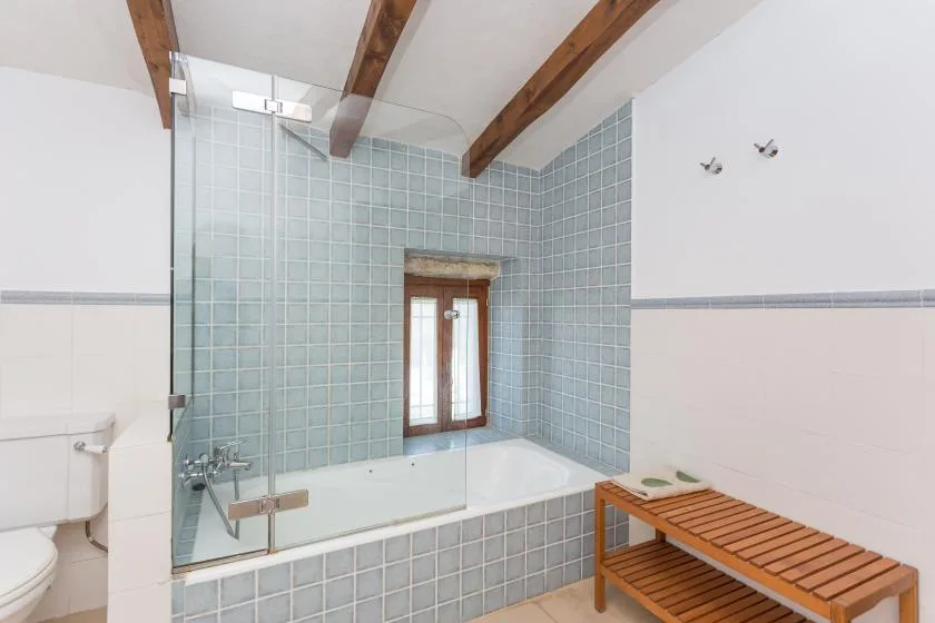 A tiled bathroom with a bathtub, glass screen, and wooden bench.