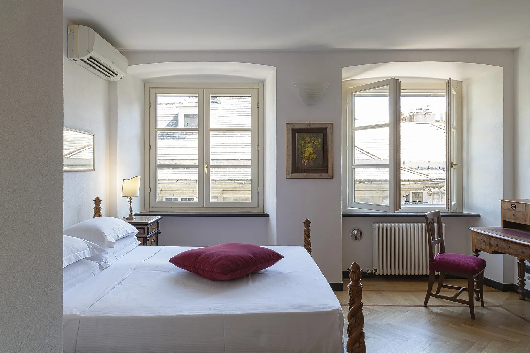 Un dormitorio luminoso con una cama y ventanas con vistas a los tejados cercanos.