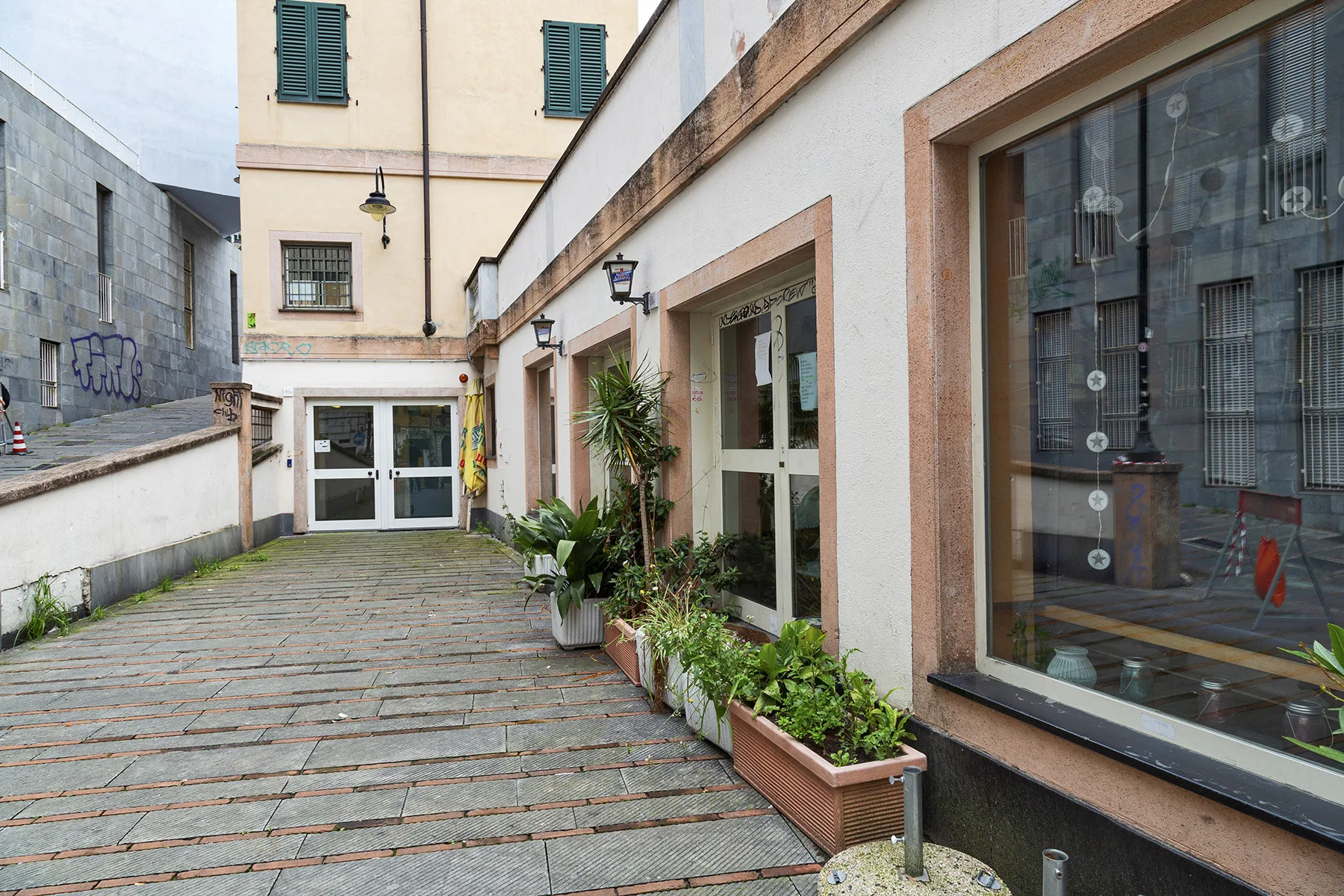 Un pasillo pavimentado de patio bordea un edificio con grandes ventanas y plantas en macetas.