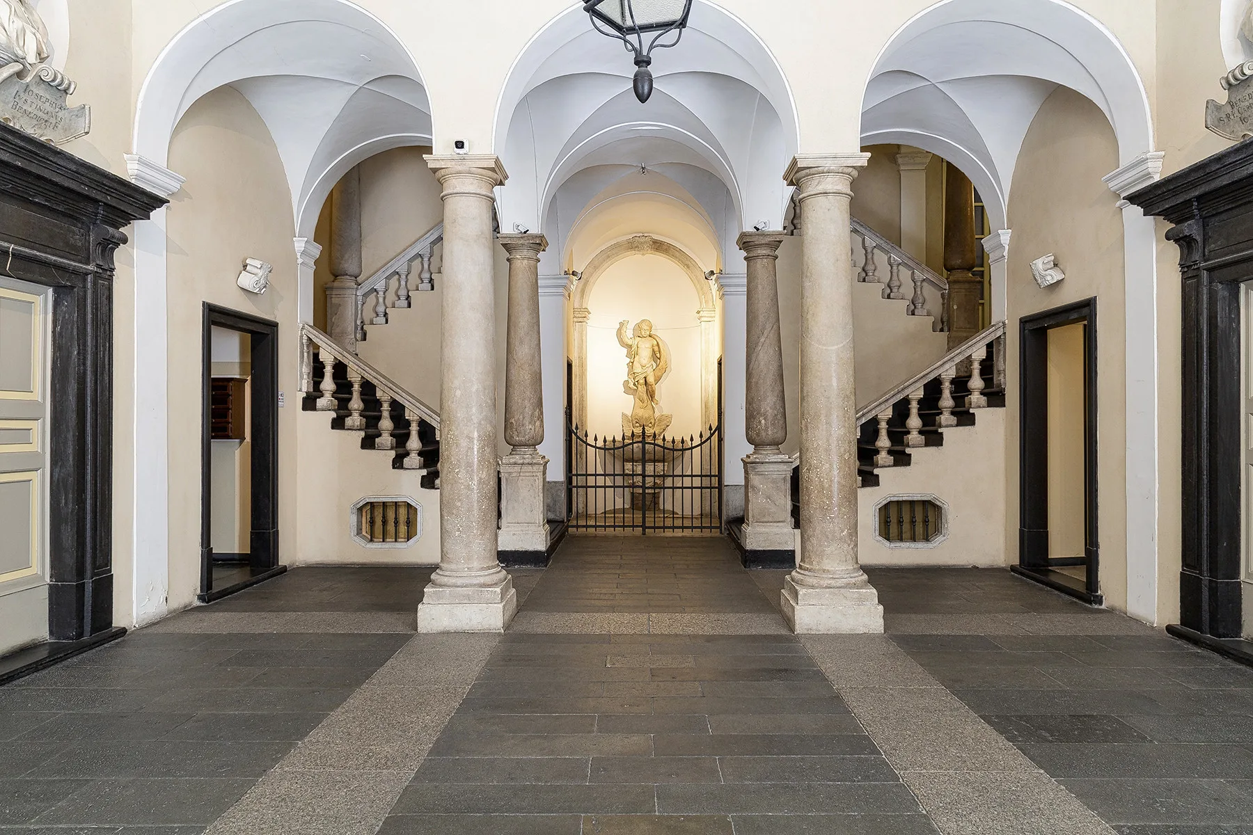 Un ornamentado vestíbulo interior con techos abovedados, escaleras y una estatua tras una reja al fondo.