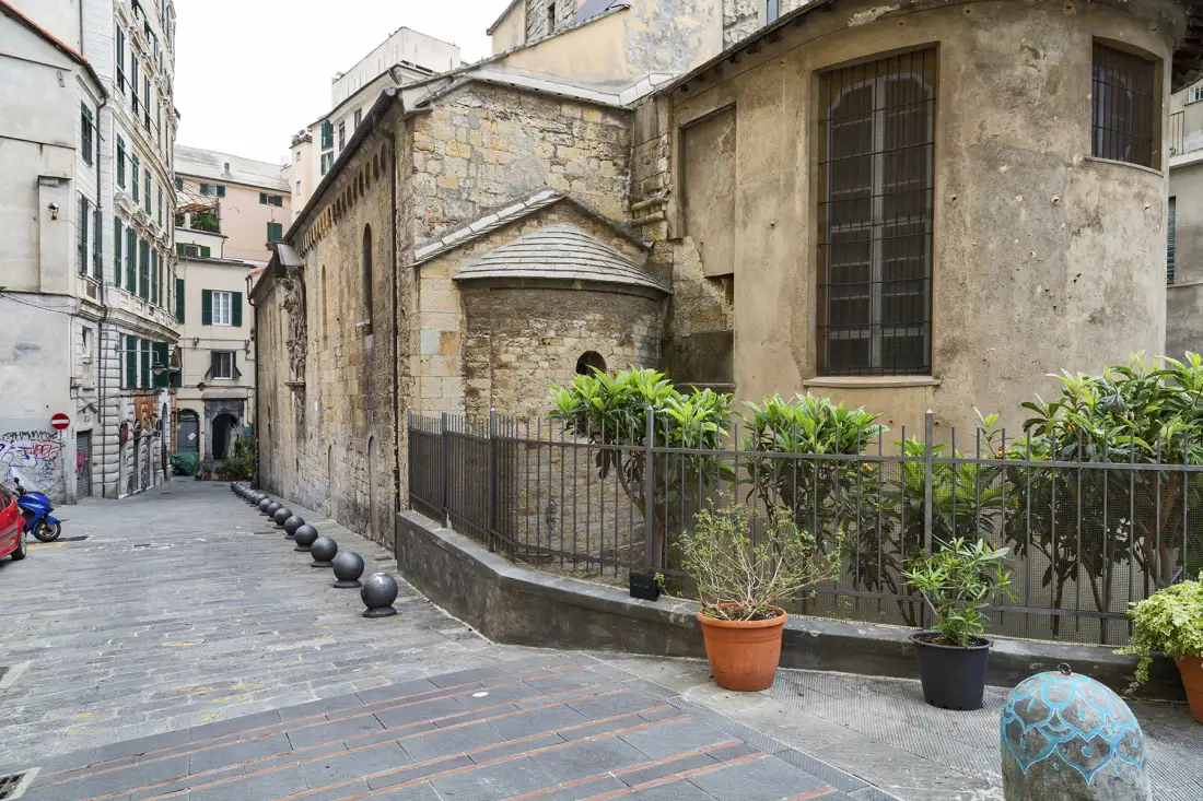 Un estrecho callejón pavimentado pasa junto a un antiguo edificio de piedra y un jardín cercado.