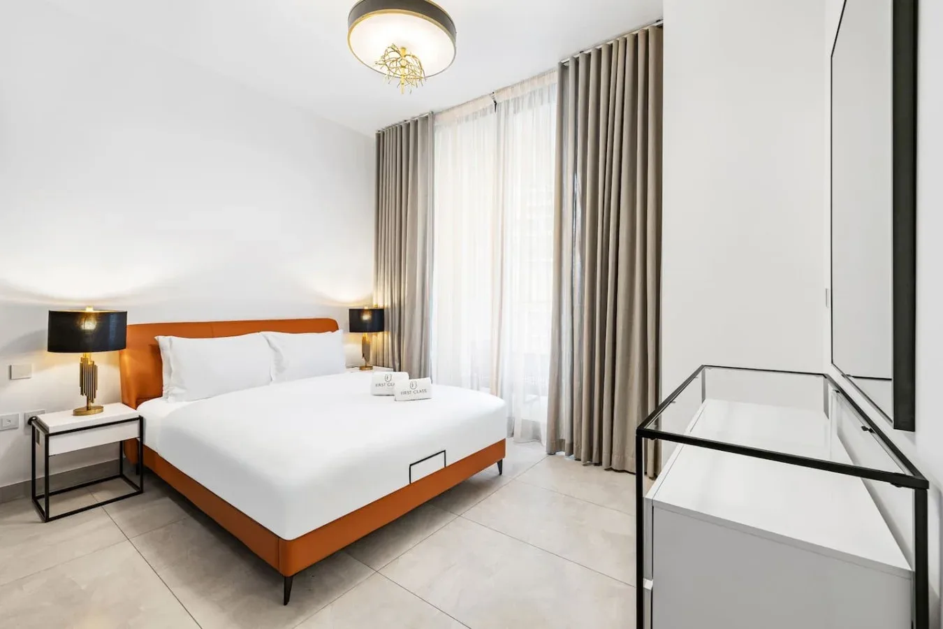 A bright bedroom with a neatly made bed and tall curtained windows.
