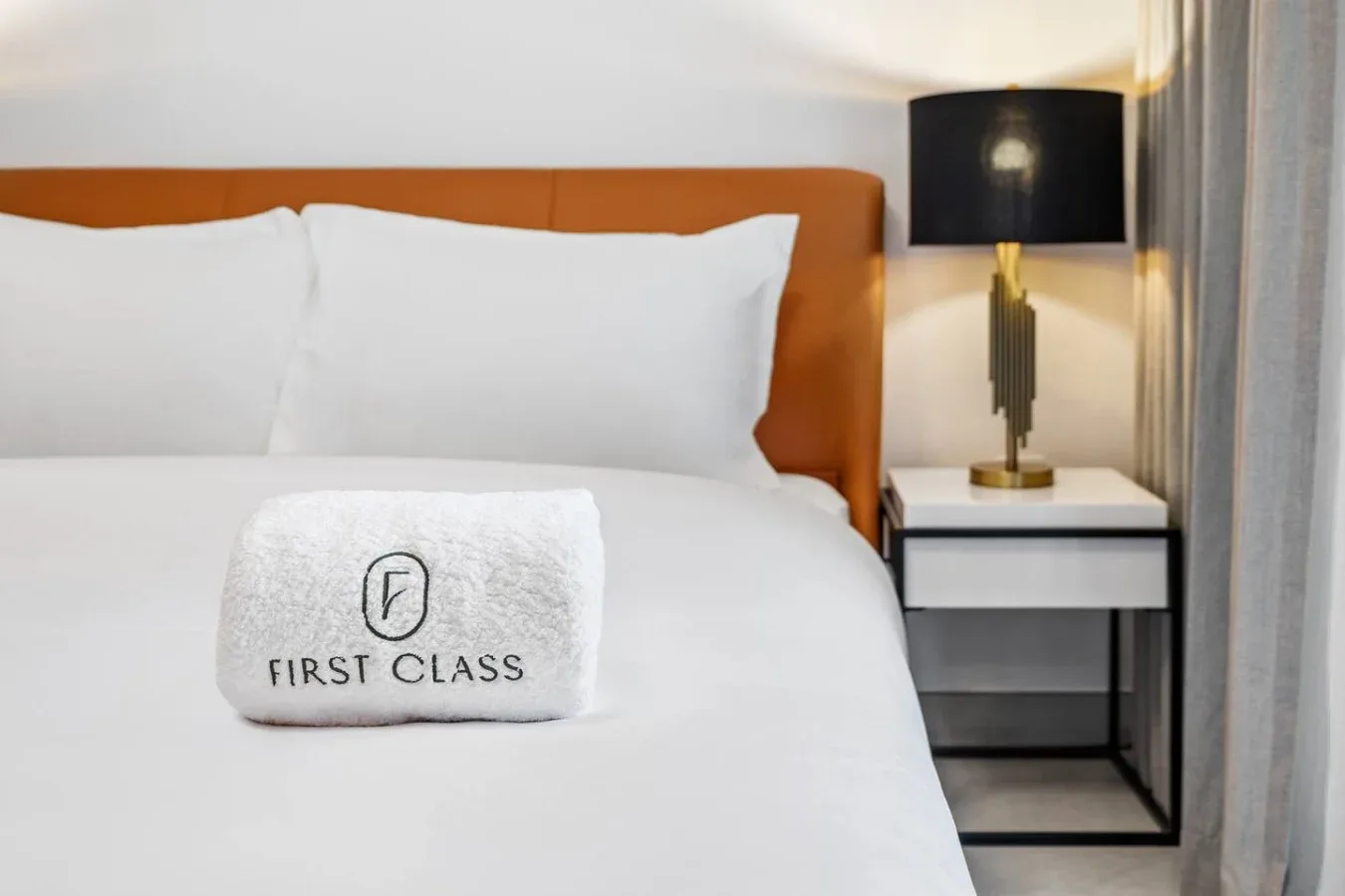 A neatly made bed with white bedding and a folded towel sits beside a lamp.
