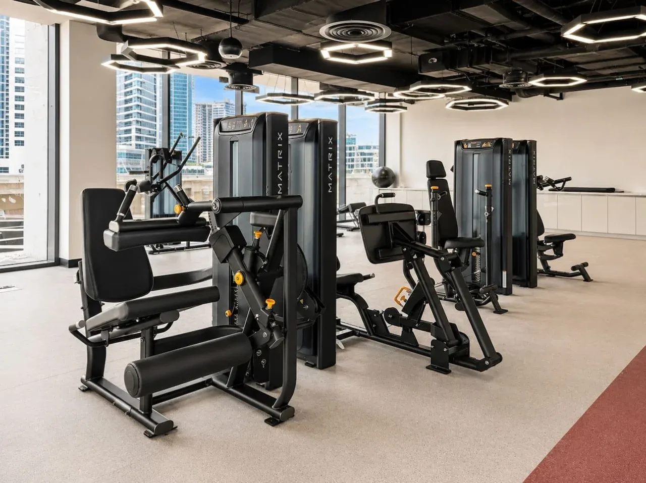 A gym with several exercise machines and large windows overlooking nearby buildings.