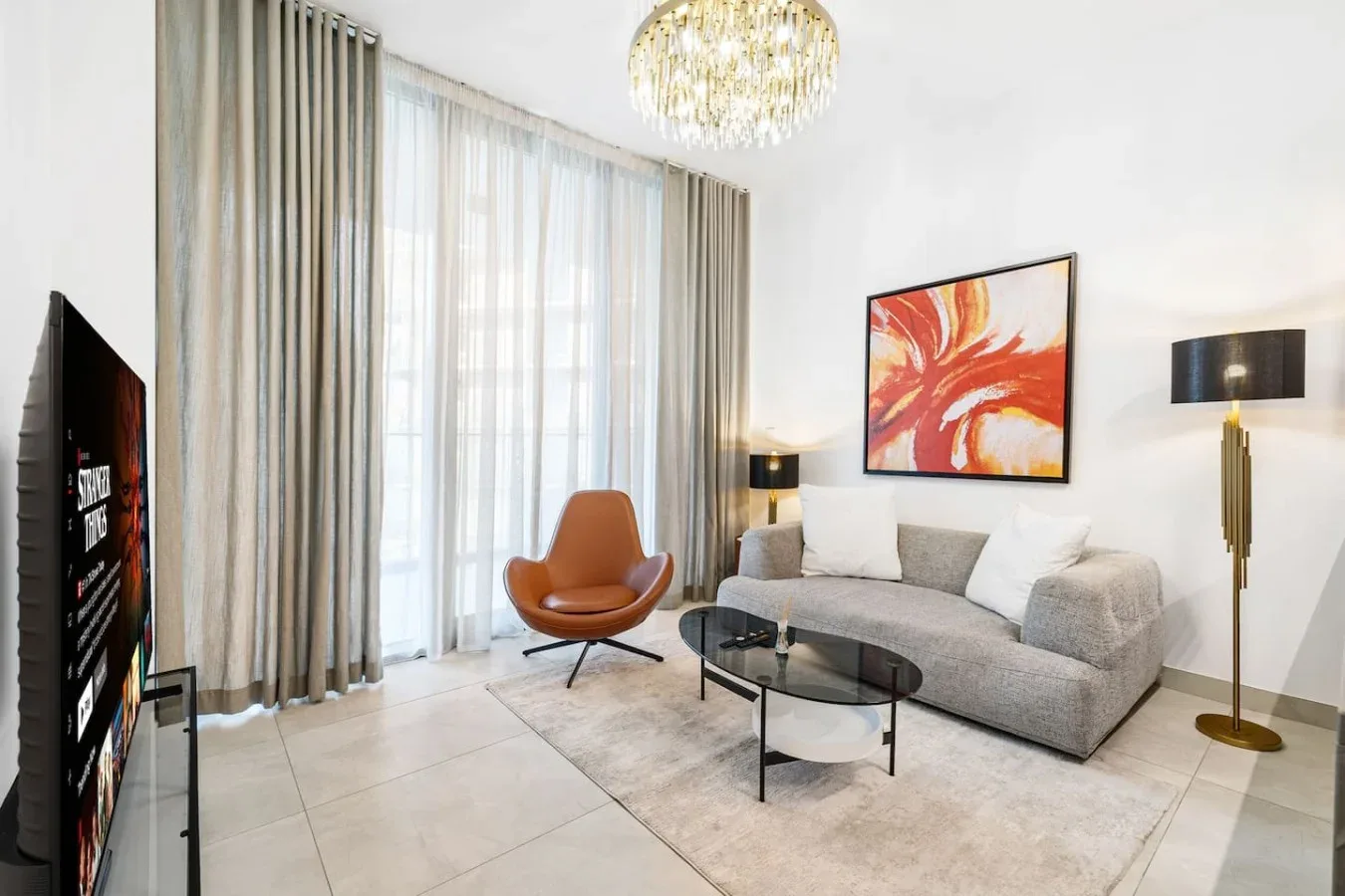 A bright living room with a gray sofa, glass coffee table, and large curtained windows.