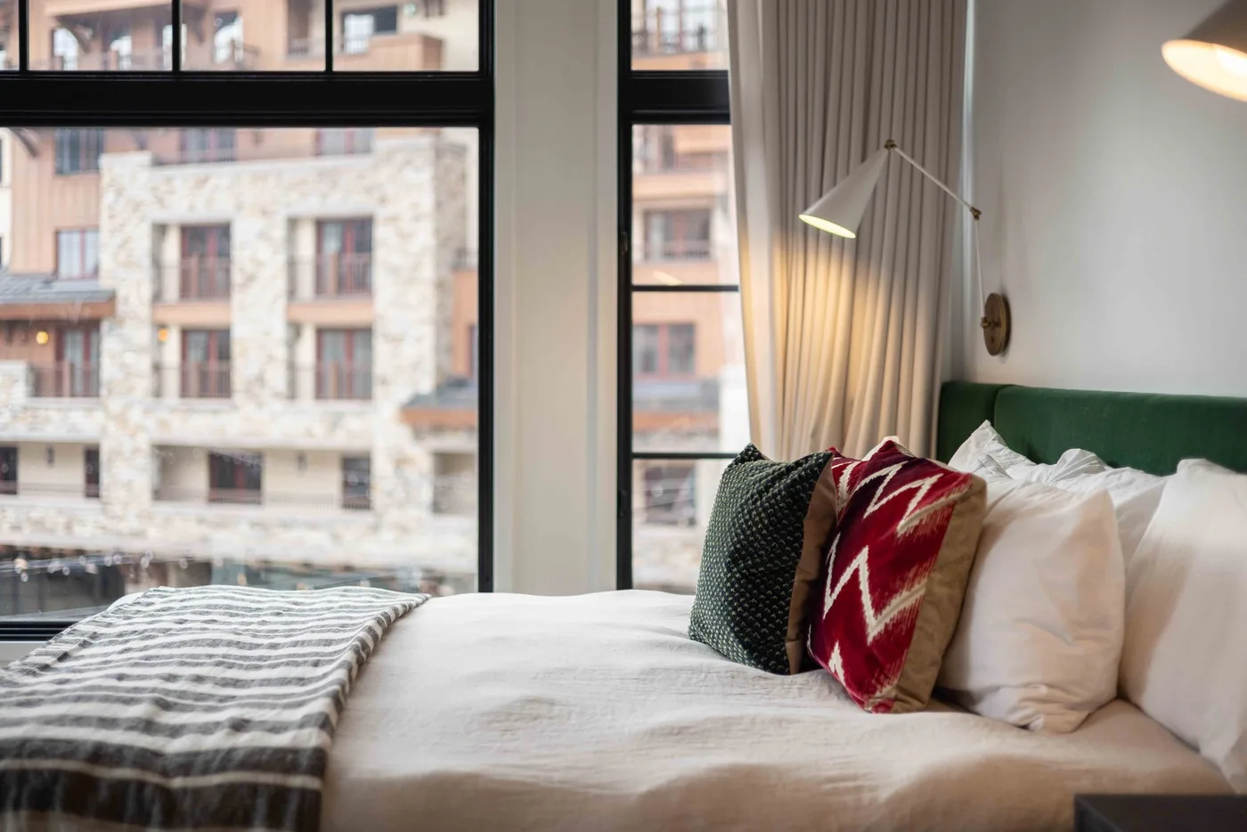 Un dormitorio con una cama tendida, cojines estampados y ventanas grandes.