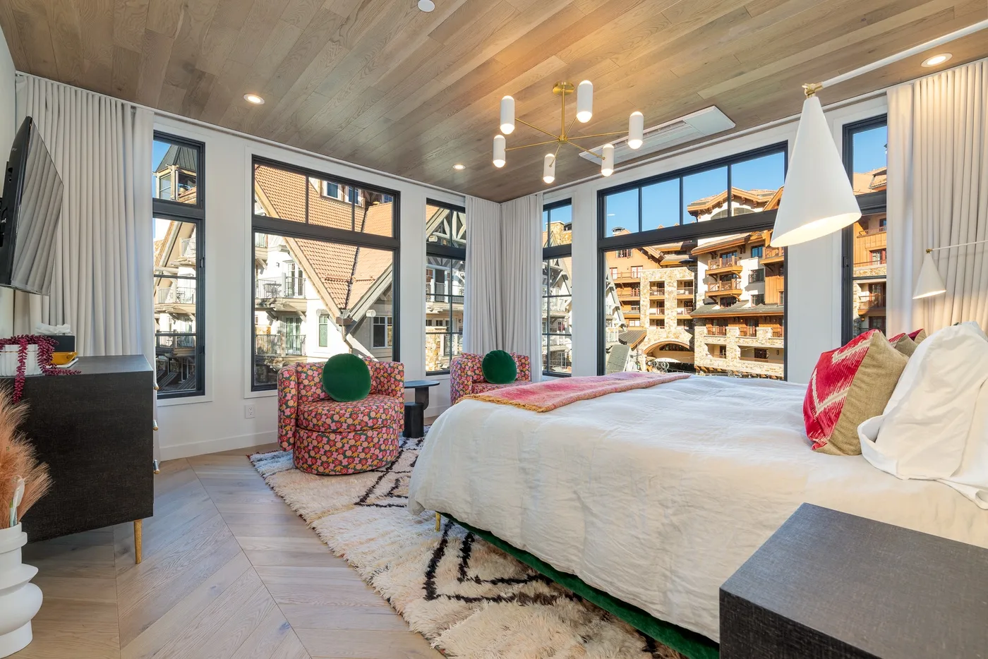 Dormitorio luminoso con una cama grande junto a amplias ventanas con vistas a edificios cercanos.