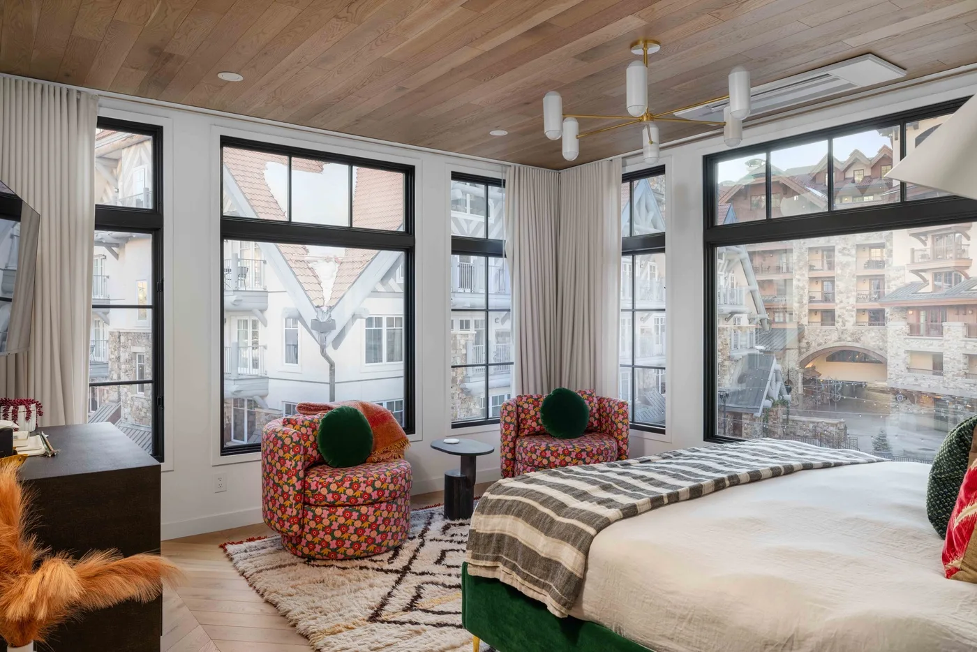 Un dormitorio luminoso tiene una cama grande, asientos coloridos y ventanas altas.