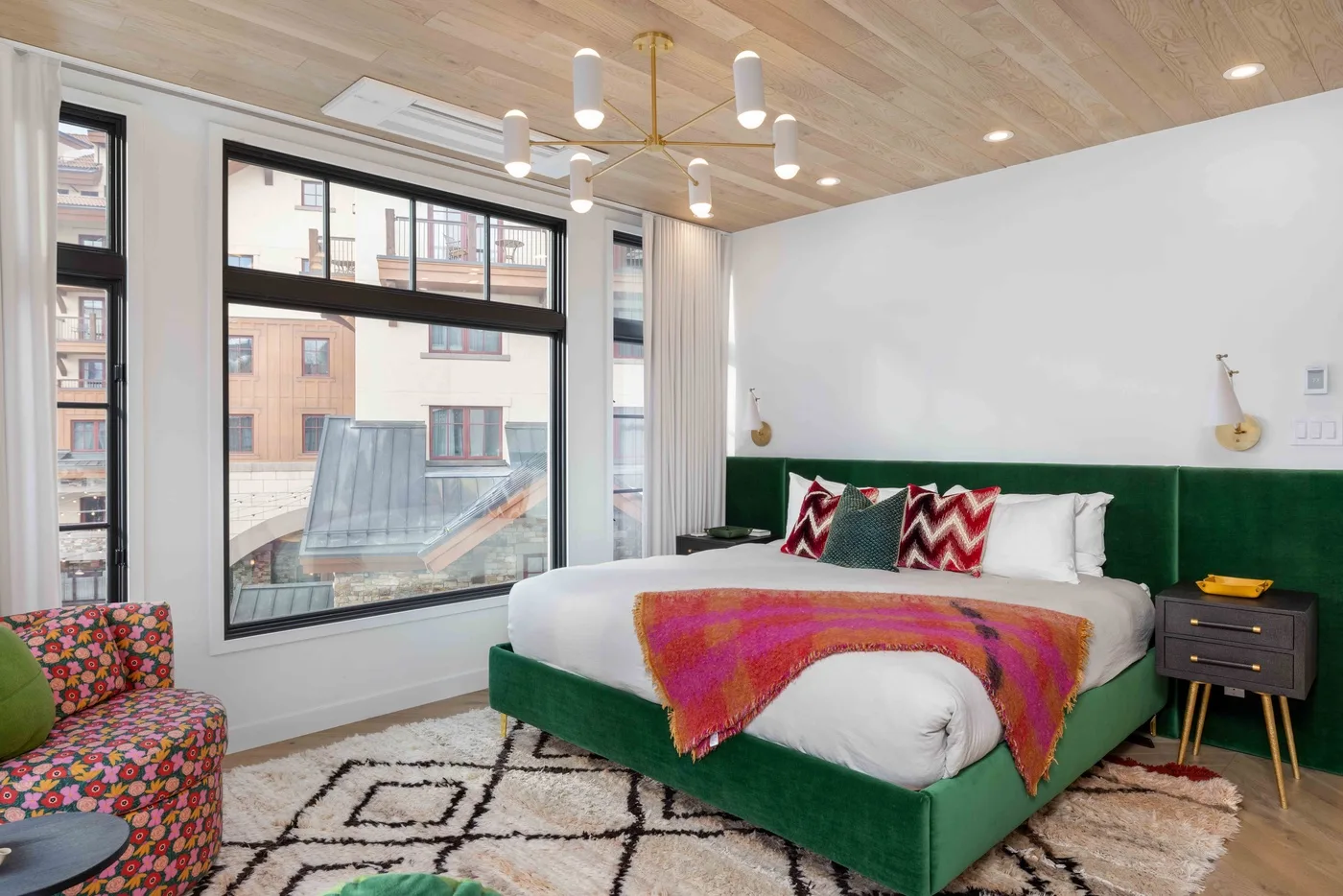Un dormitorio con una cama verde, ropa de cama colorida y ventanas grandes.