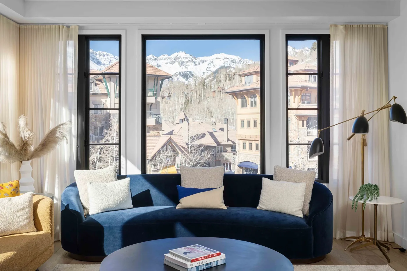 Una sala luminosa con un sofá azul frente a grandes ventanas y vistas a las montañas.