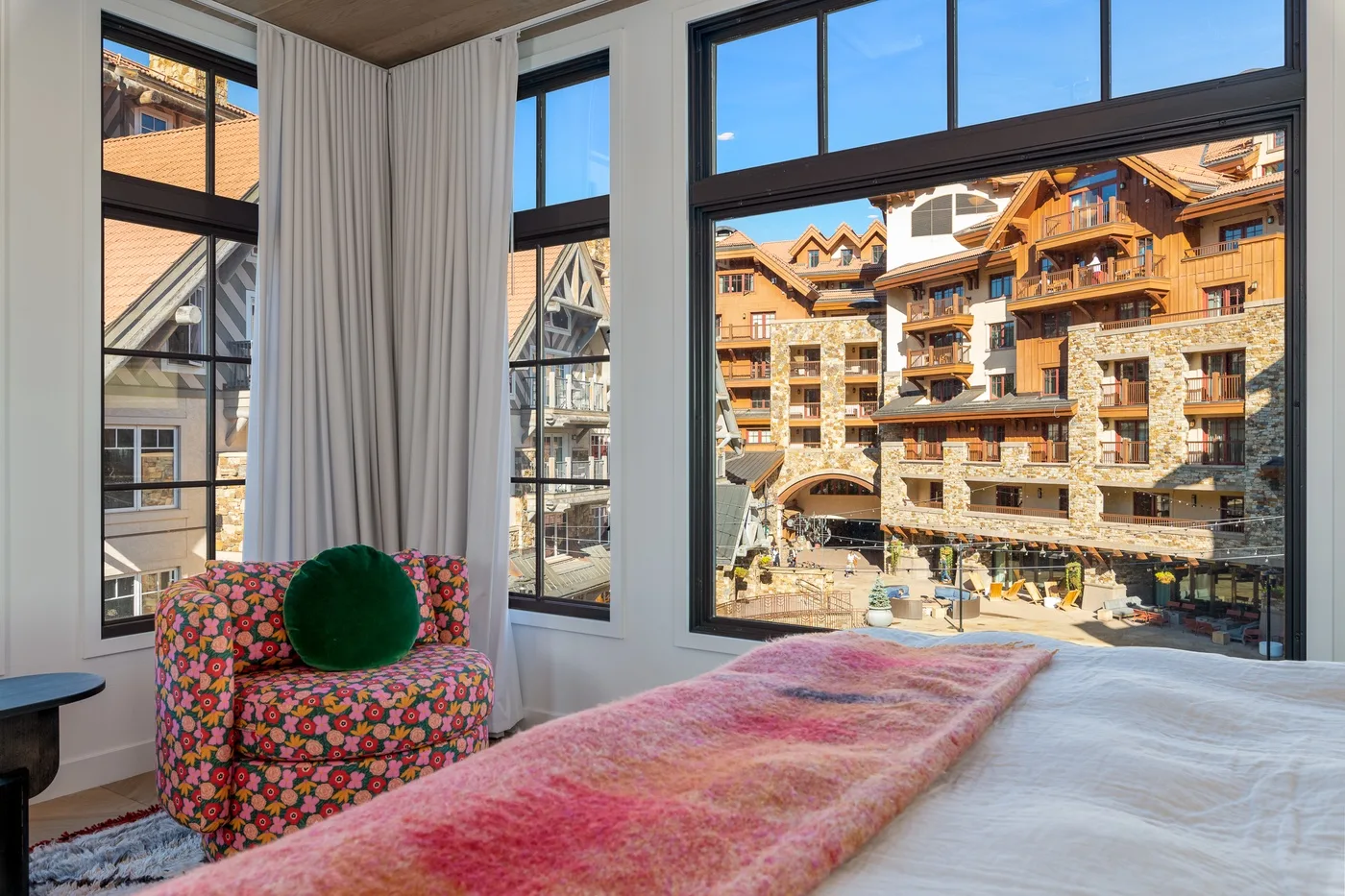 Un dormitorio con una gran ventana con vistas a un edificio de estilo alpino iluminado por el sol.