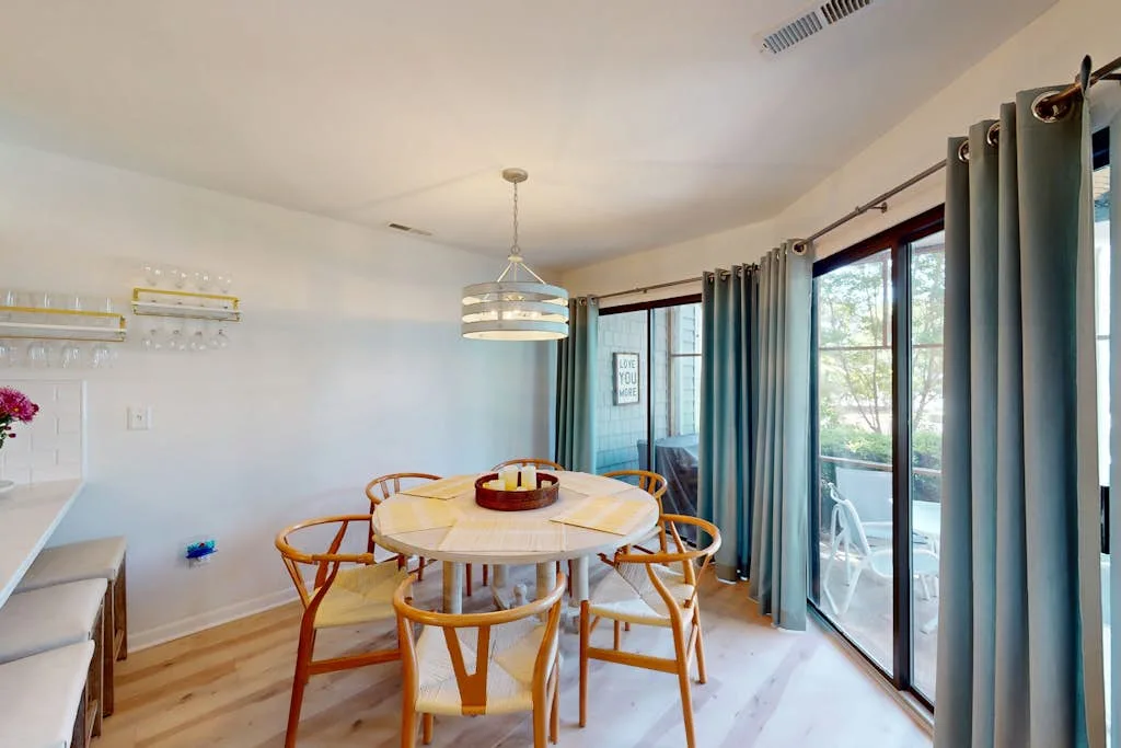 A dining area with a round table and seating beside large curtained windows.