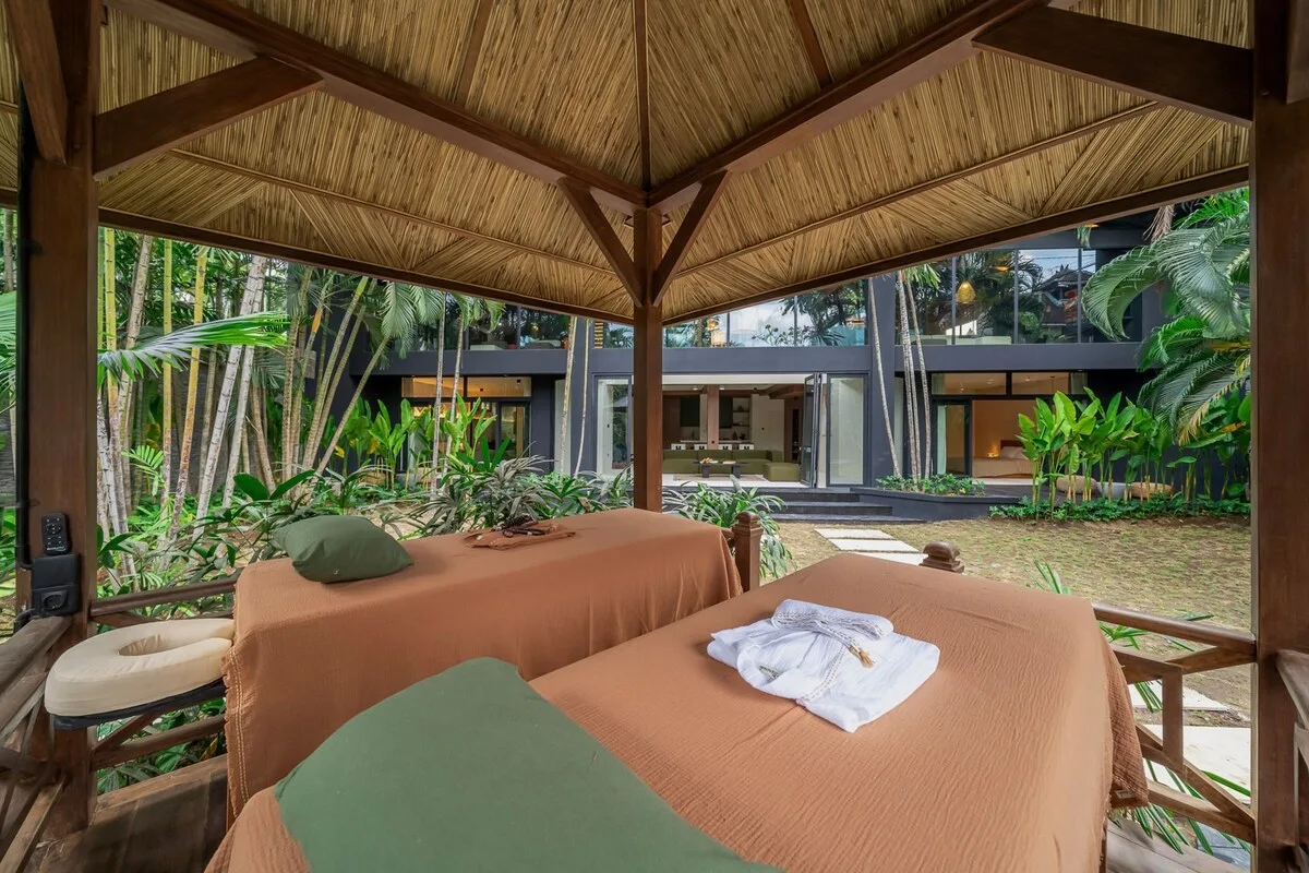 An open-air seating area beneath a thatched pavilion overlooks a lush garden and modern building.