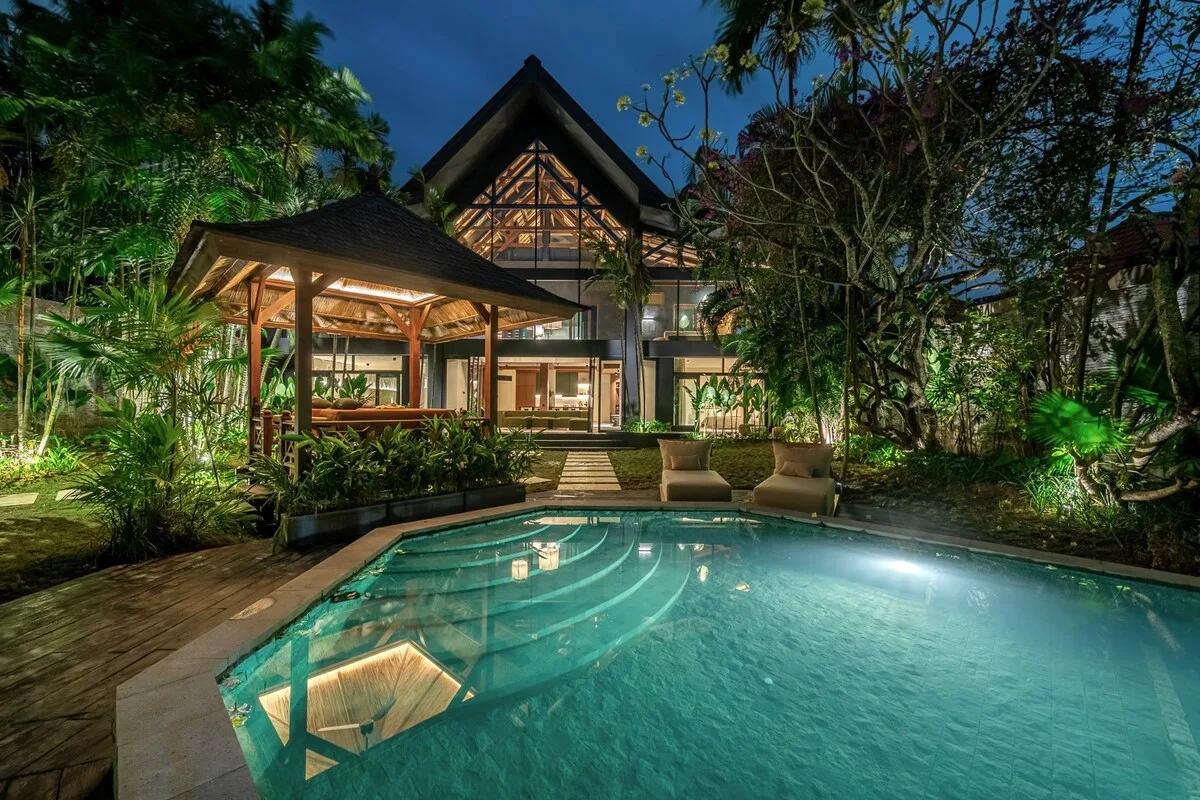 A swimming pool sits in front of a warmly lit house surrounded by tropical greenery.