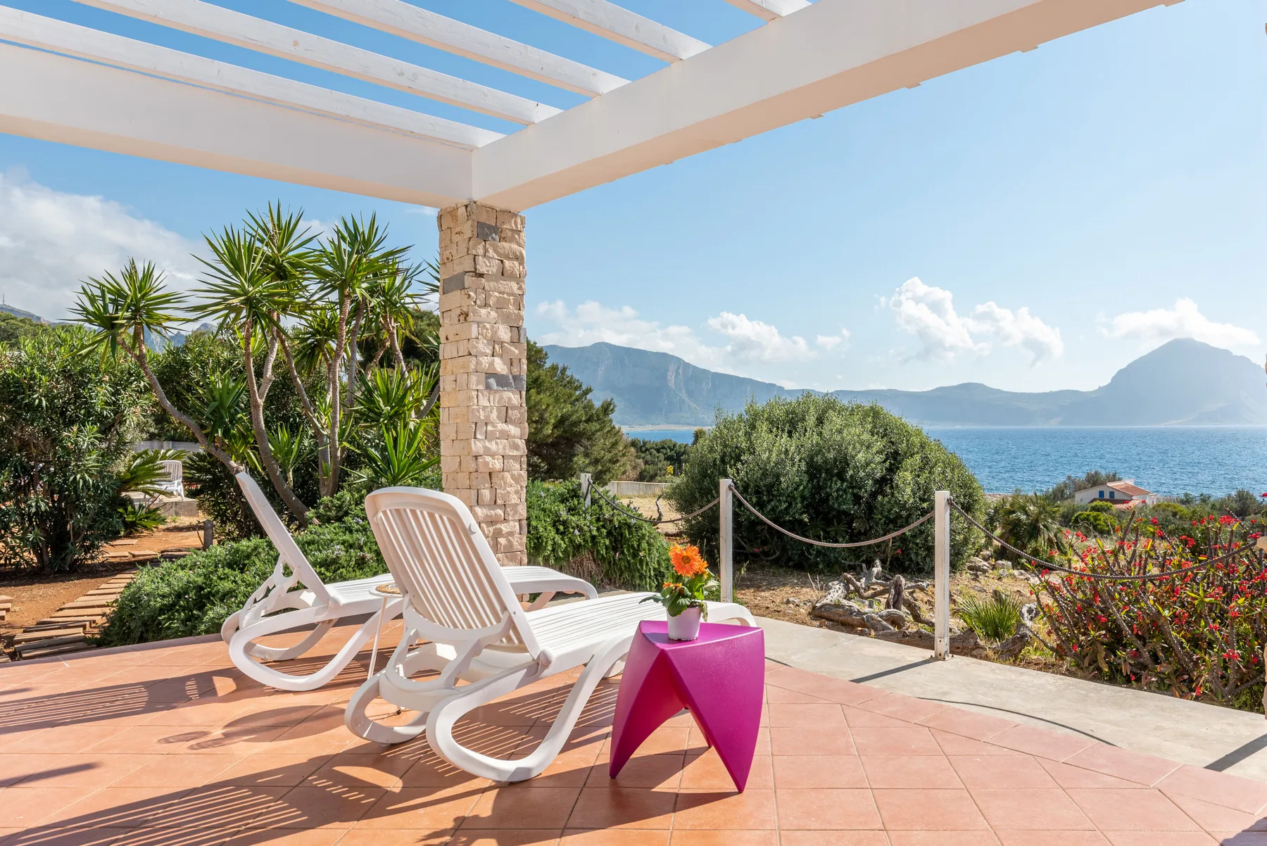 Una terraza soleada al aire libre tiene asientos y vistas al mar y las montañas.