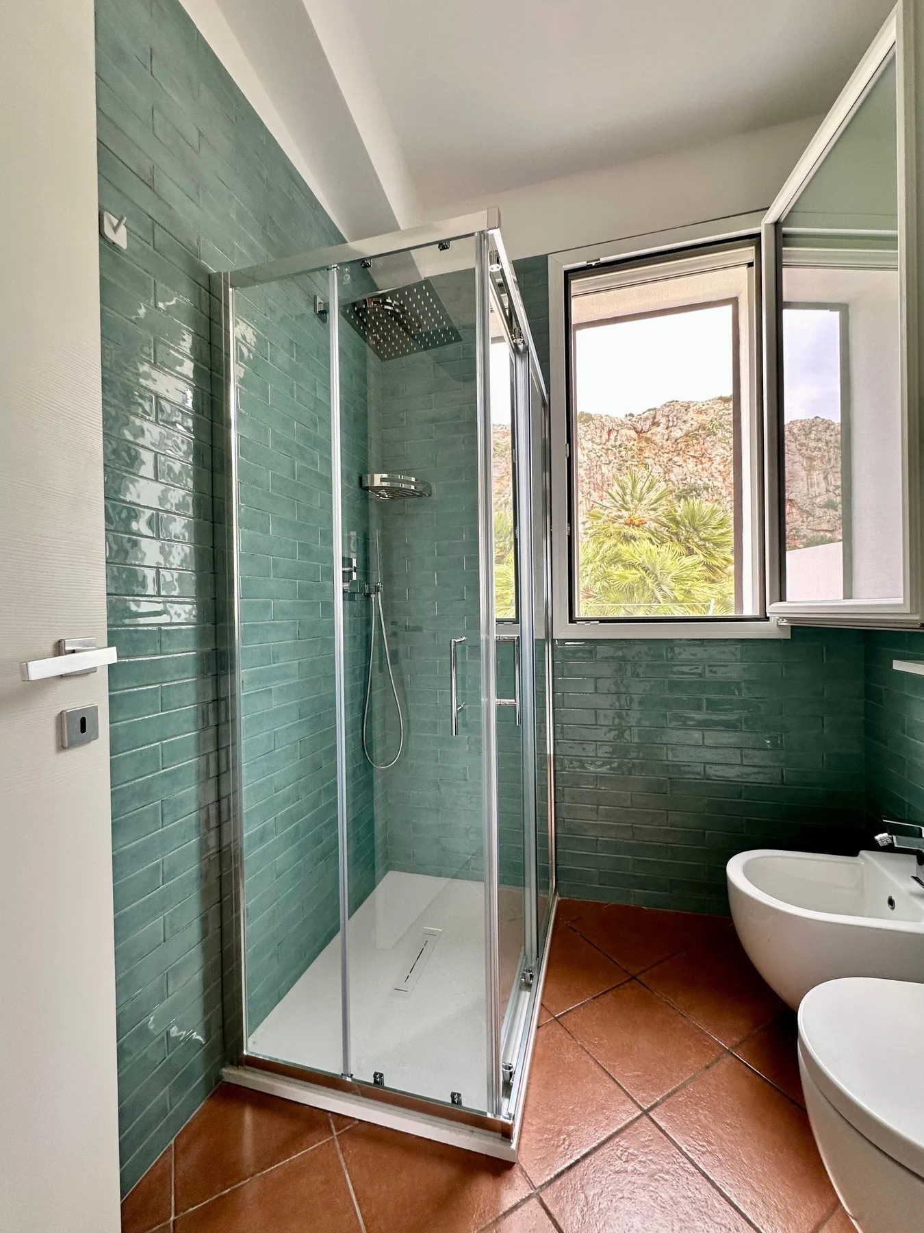 Un baño con ducha de vidrio, paredes de azulejos verdes y una ventana.