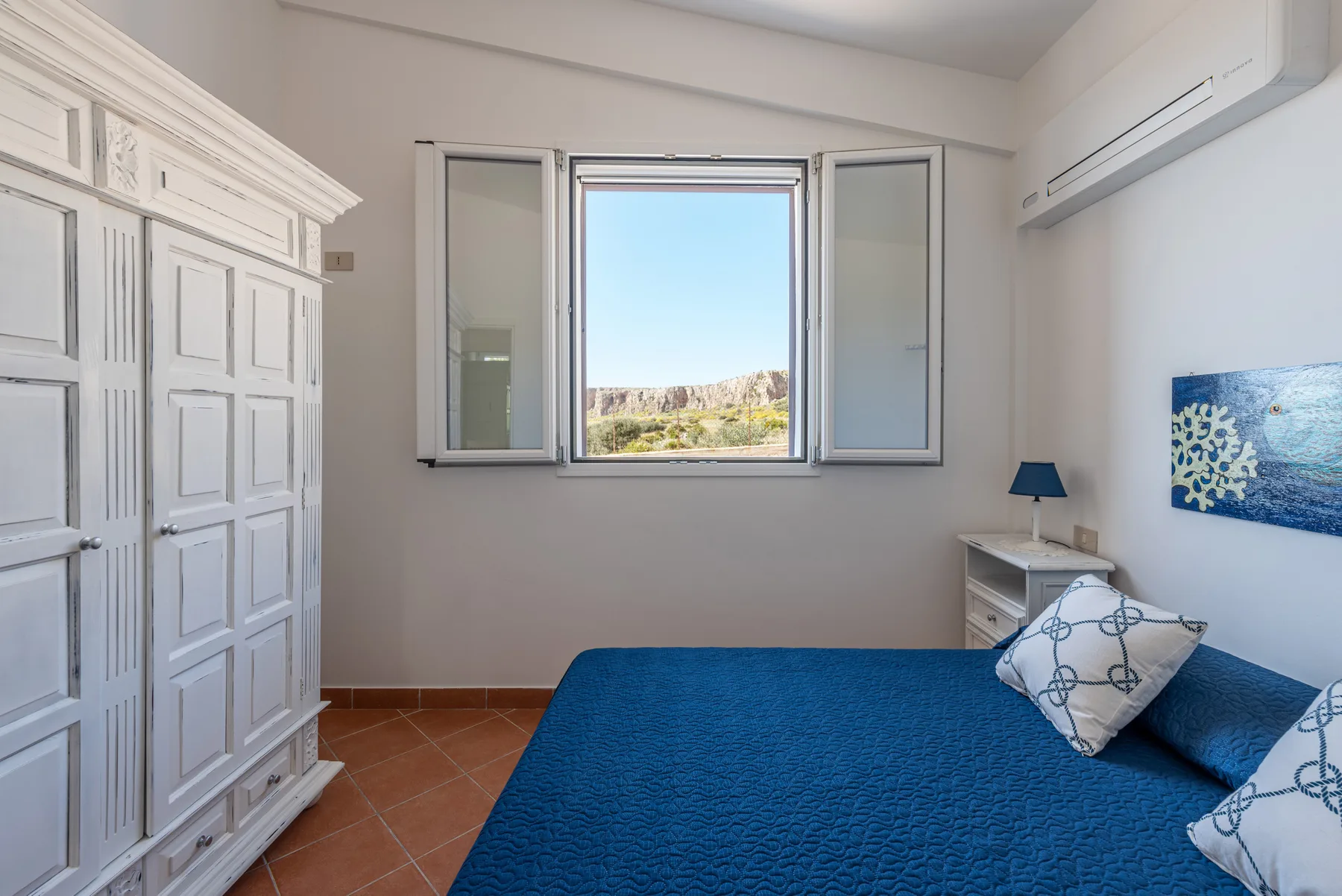 Dormitorio con una colcha azul y una ventana abierta con vistas al paisaje.