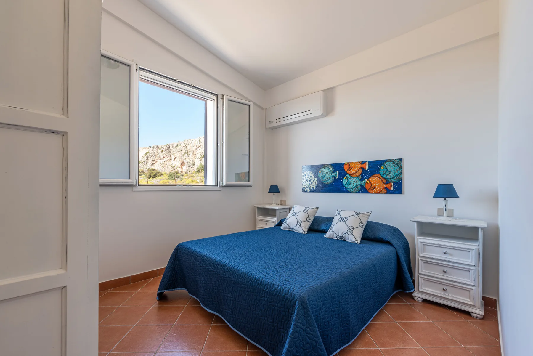 Un dormitorio con una colcha azul, mesitas de noche y una ventana.