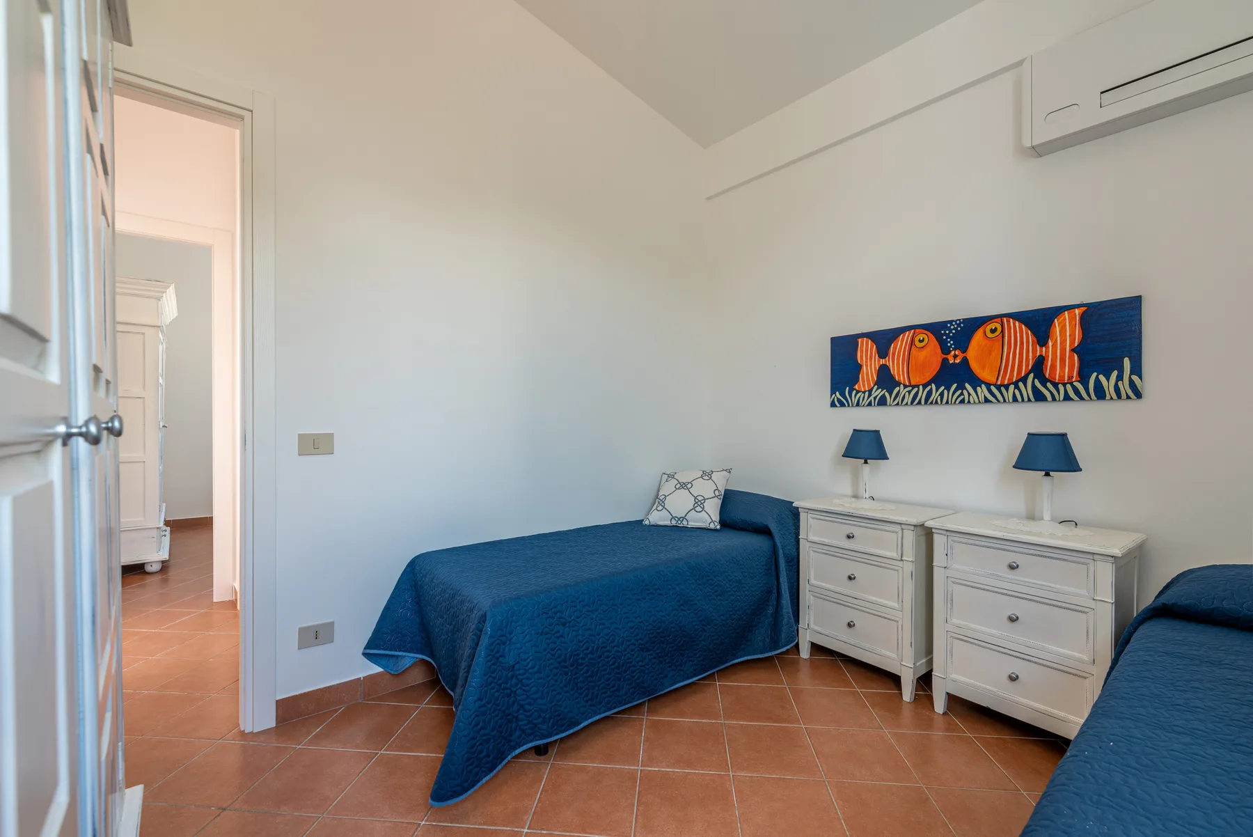 Un dormitorio con camas cubiertas de azul, cómodas blancas y un colorido cuadro de peces.