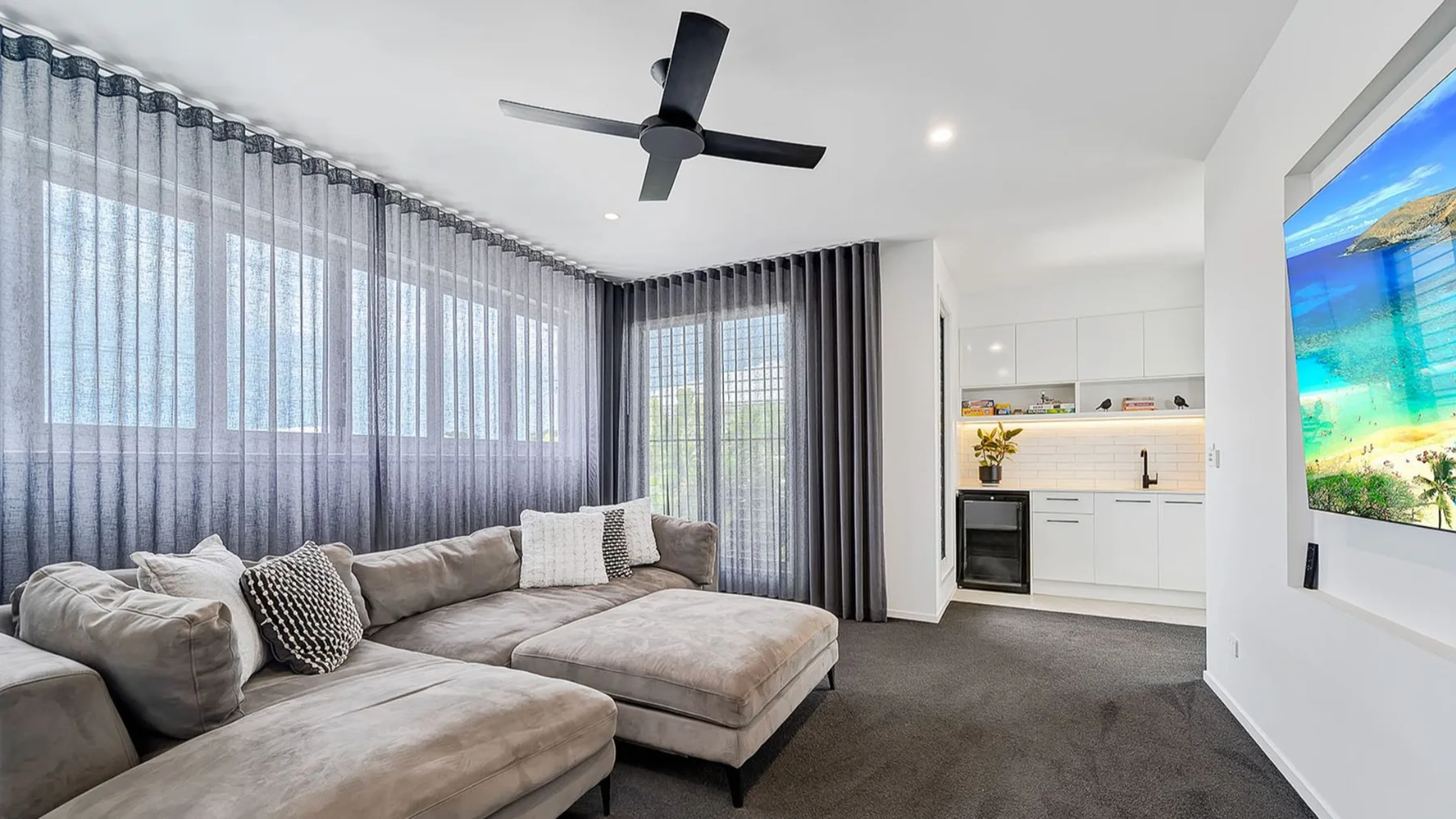 A bright living room with seating, floor-to-ceiling curtains, and a wall-mounted television.