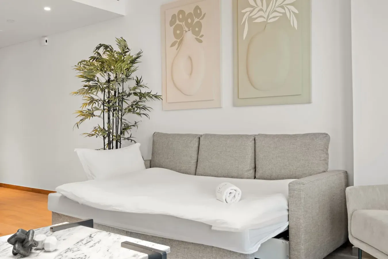 Una sala luminosa con un sofá gris, ropa de cama blanca y láminas botánicas enmarcadas.