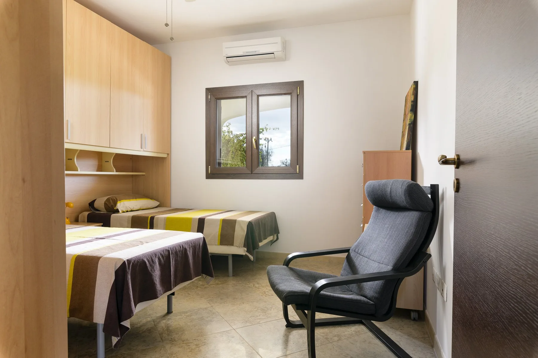 Un dormitorio con dos camas y una silla tapizada oscura junto a la ventana.