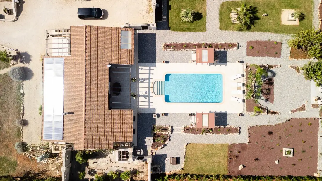 Una vista aérea muestra una piscina rectangular junto a un edificio con techo de tejas y jardines.