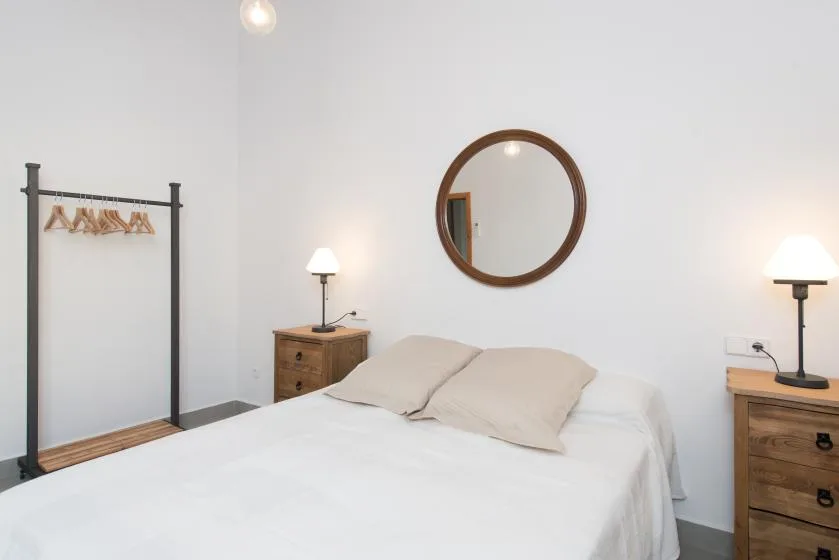 Un dormitorio luminoso con una cama, mesitas de madera y un espejo redondo en la pared.