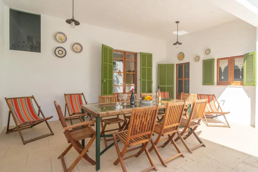 Un comedor luminoso con una mesa larga y asientos de madera junto a ventanas con contraventanas.