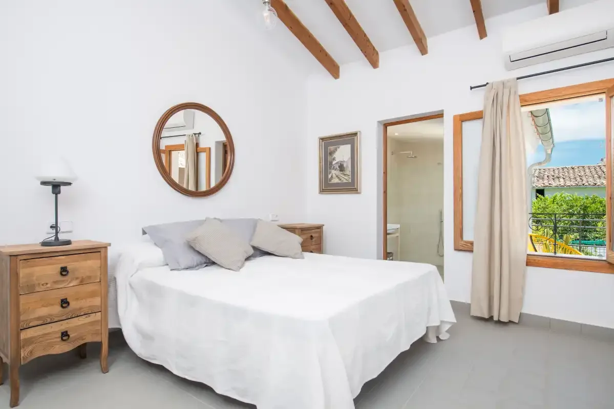 Un dormitorio luminoso con una cama blanca, vigas de madera y una ventana con vistas a la vegetación.