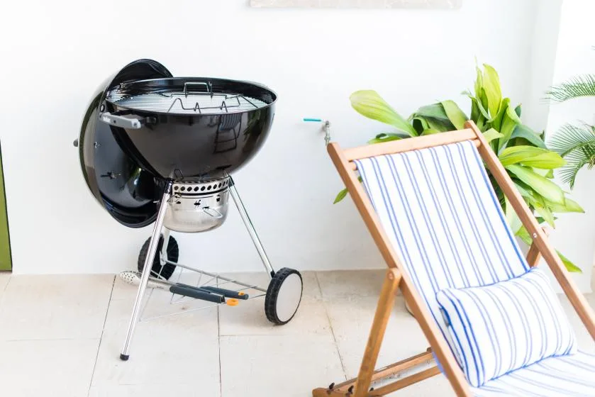 Una parrilla negra está junto a una silla plegable a rayas y plantas frondosas.