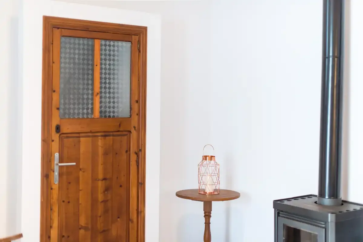 Una puerta de madera y una mesa pequeña están junto a una estufa negra en una habitación luminosa.