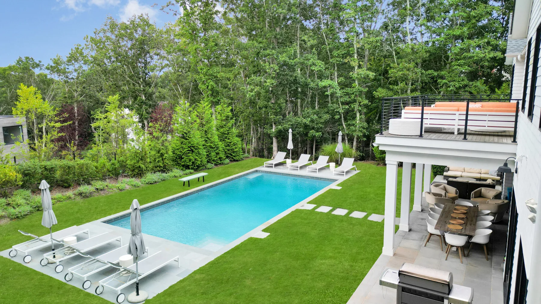 Una piscina exterior junto a un césped, con asientos en un patio cubierto.