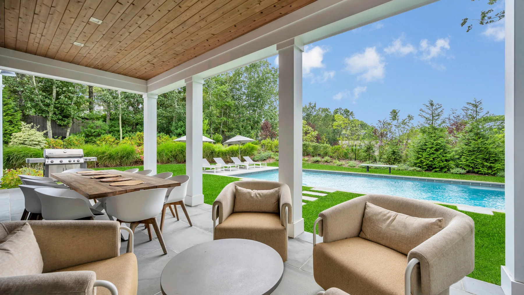 Un patio cubierto con asientos da a una piscina y un jardín.