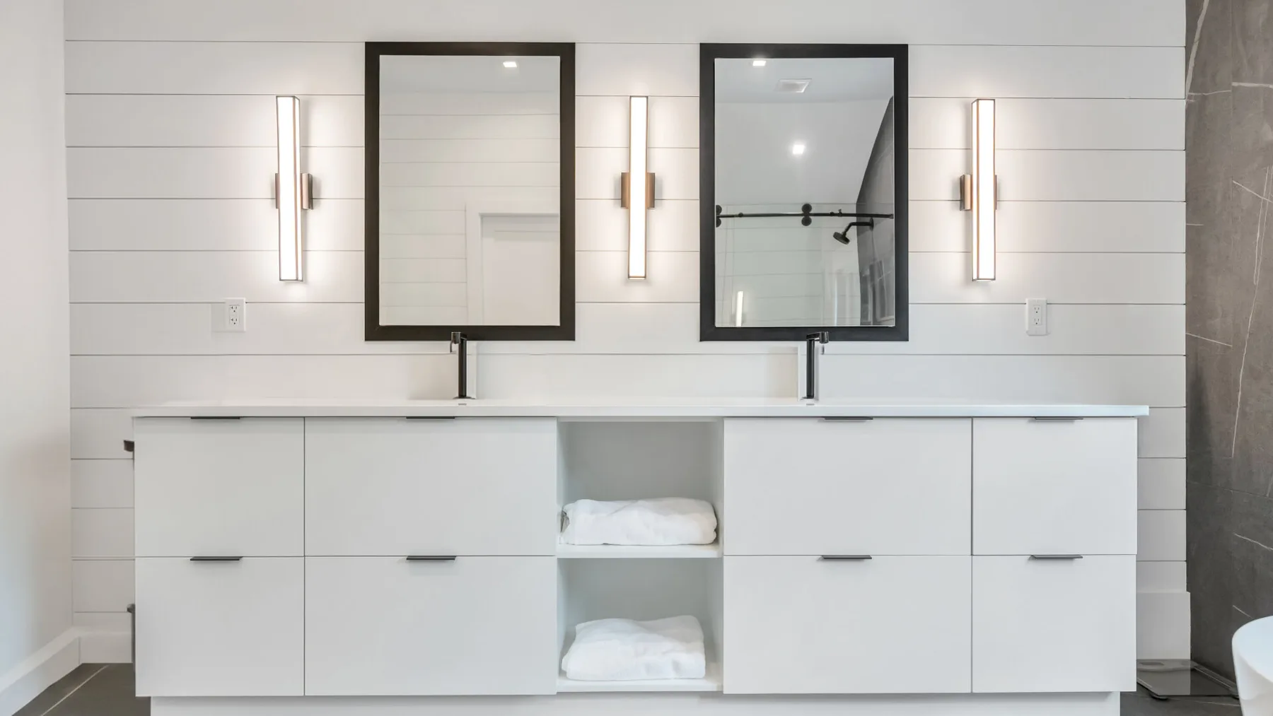 Un tocador de baño luminoso con dos espejos y toallas dobladas en estantes abiertos.