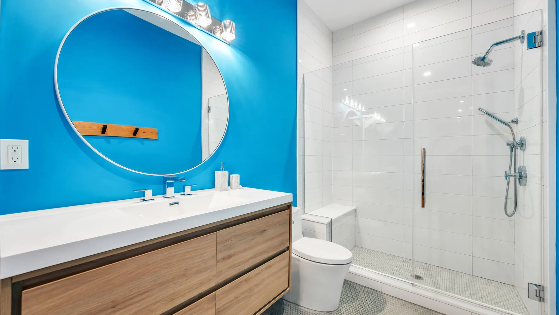 Un baño luminoso con tocador de madera, espejo redondo, inodoro y ducha de vidrio.