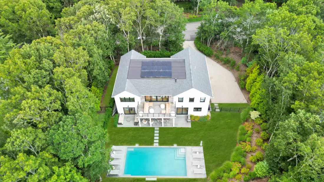 Vista aérea de una casa blanca, césped y piscina rodeados de árboles.