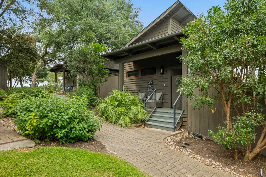 A modern house with a wooden facade is surrounded by lush greenery and a stone-paved walkway, under a clear blue sky.