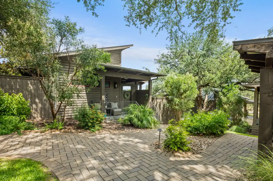 A modern house with a wooden facade is surrounded by lush greenery and a stone-paved walkway, under a clear blue sky.