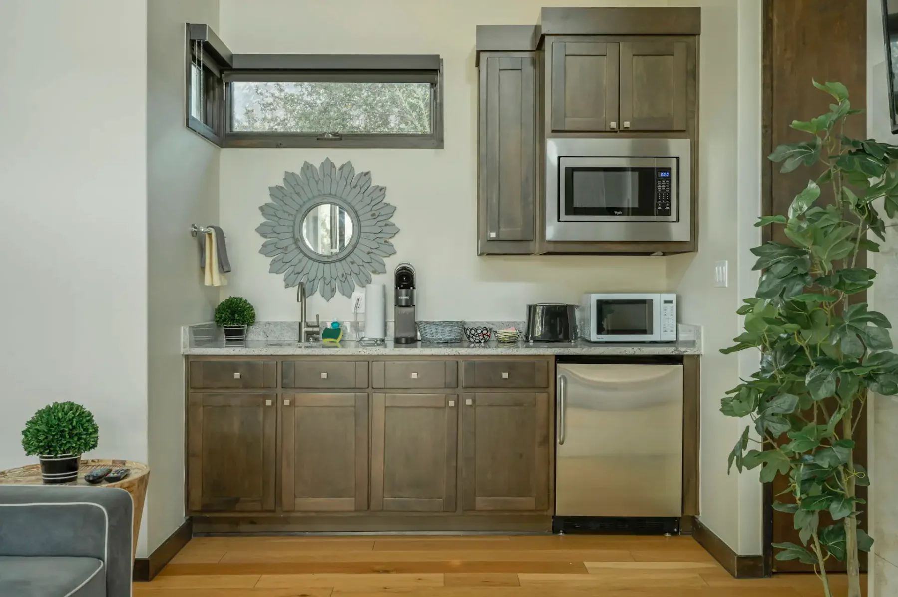 Compact kitchen with wood cabinets, microwave, mini-fridge, coffee maker, and decorative mirror. Small plants add a touch of greenery.