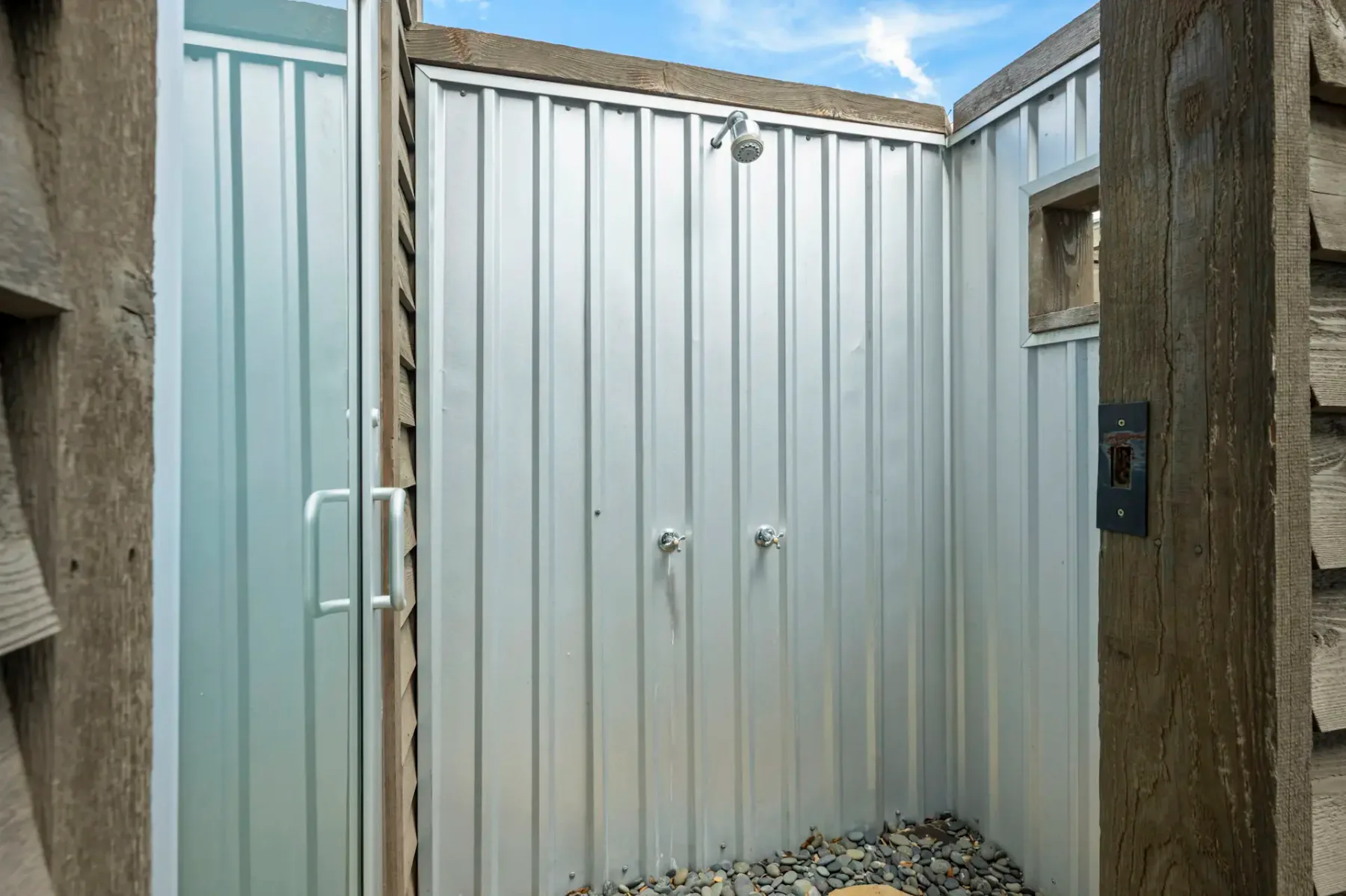 Outdoor shower with metal walls, wooden accents, and a stone floor, under a blue sky.