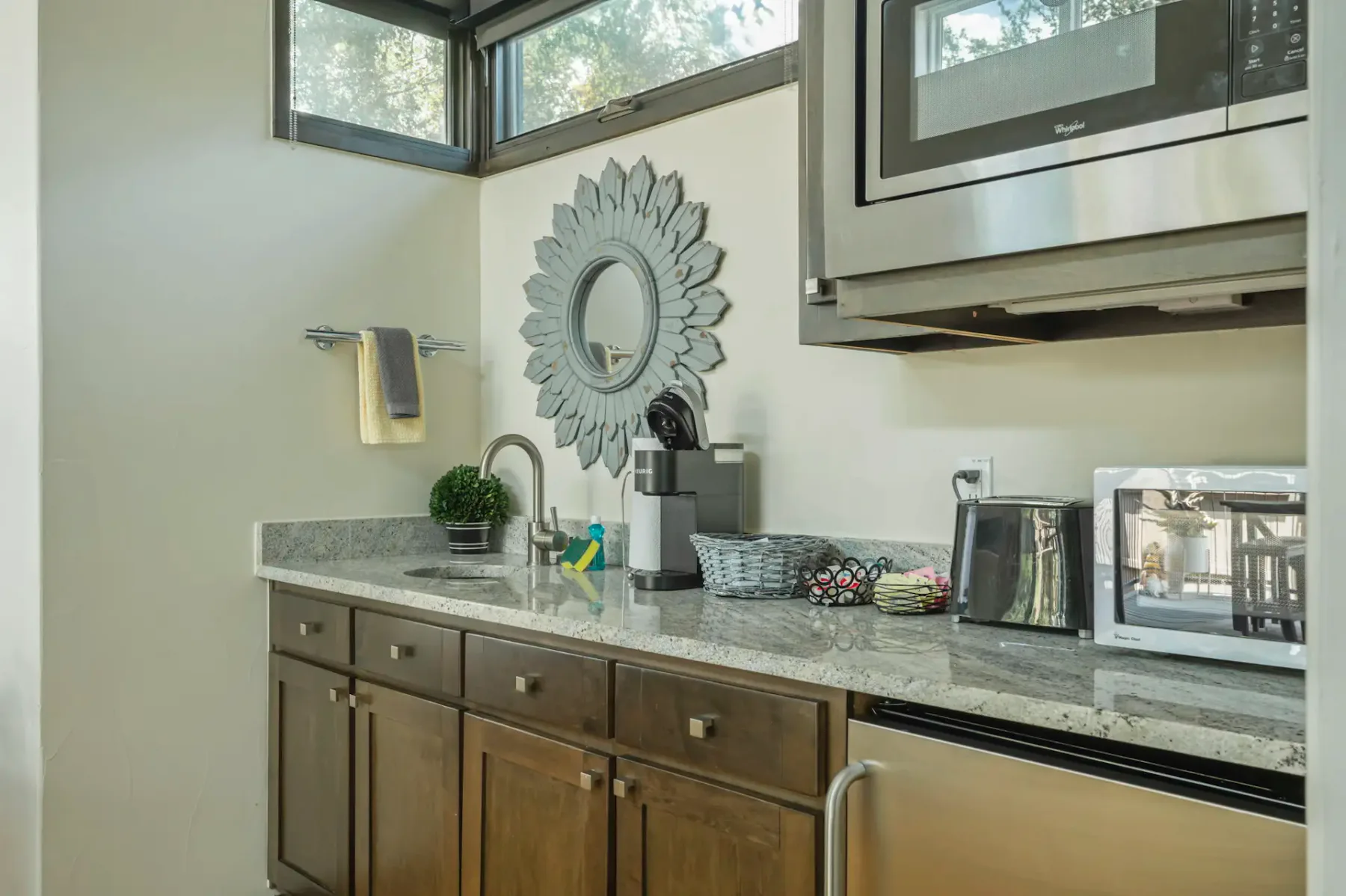 Compact kitchen with wood cabinets, microwave, mini-fridge, coffee maker, and decorative mirror. Small plants add a touch of greenery.