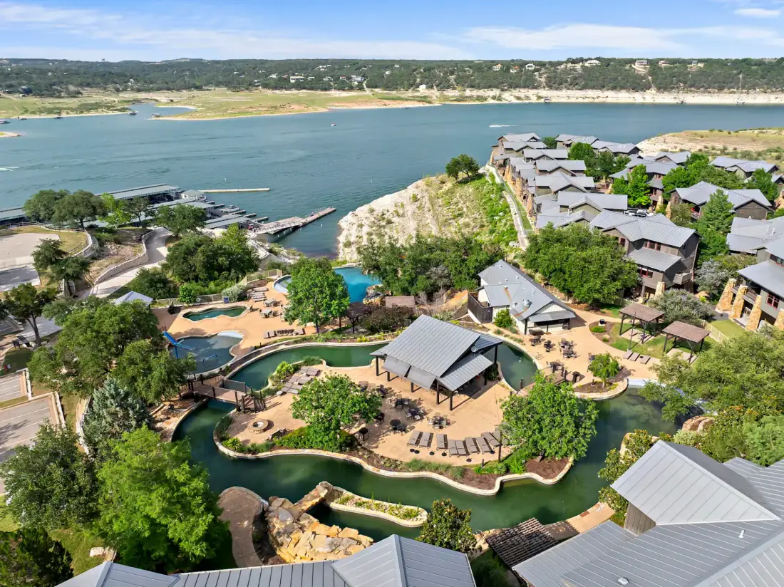 Aerial view of a residential area with houses, lush greenery, a winding pool with a water slide, and a lake in the background under a clear sky.