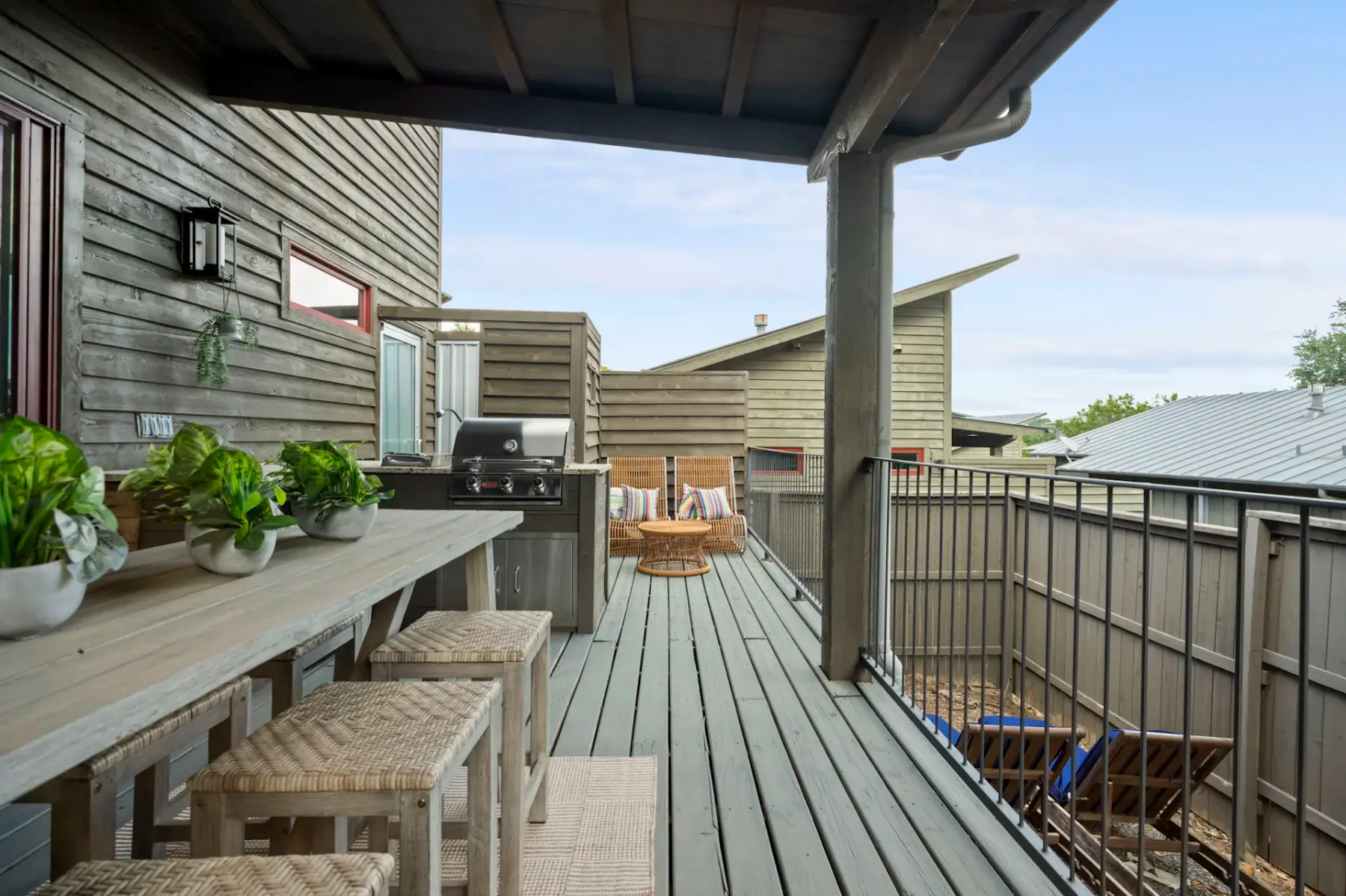 A covered wooden deck has a long counter with stools, potted plants, and a grill.
