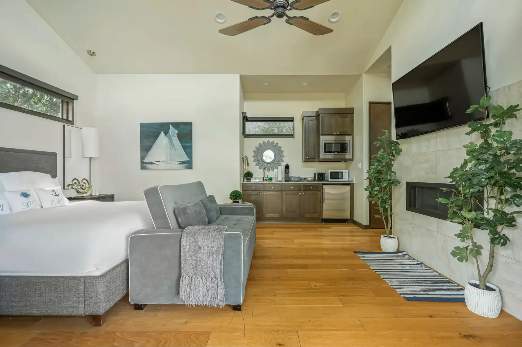 Wall-mounted TV, kitchenette, indoor plants, and wooden flooring. Sailboat artwork on the wall.