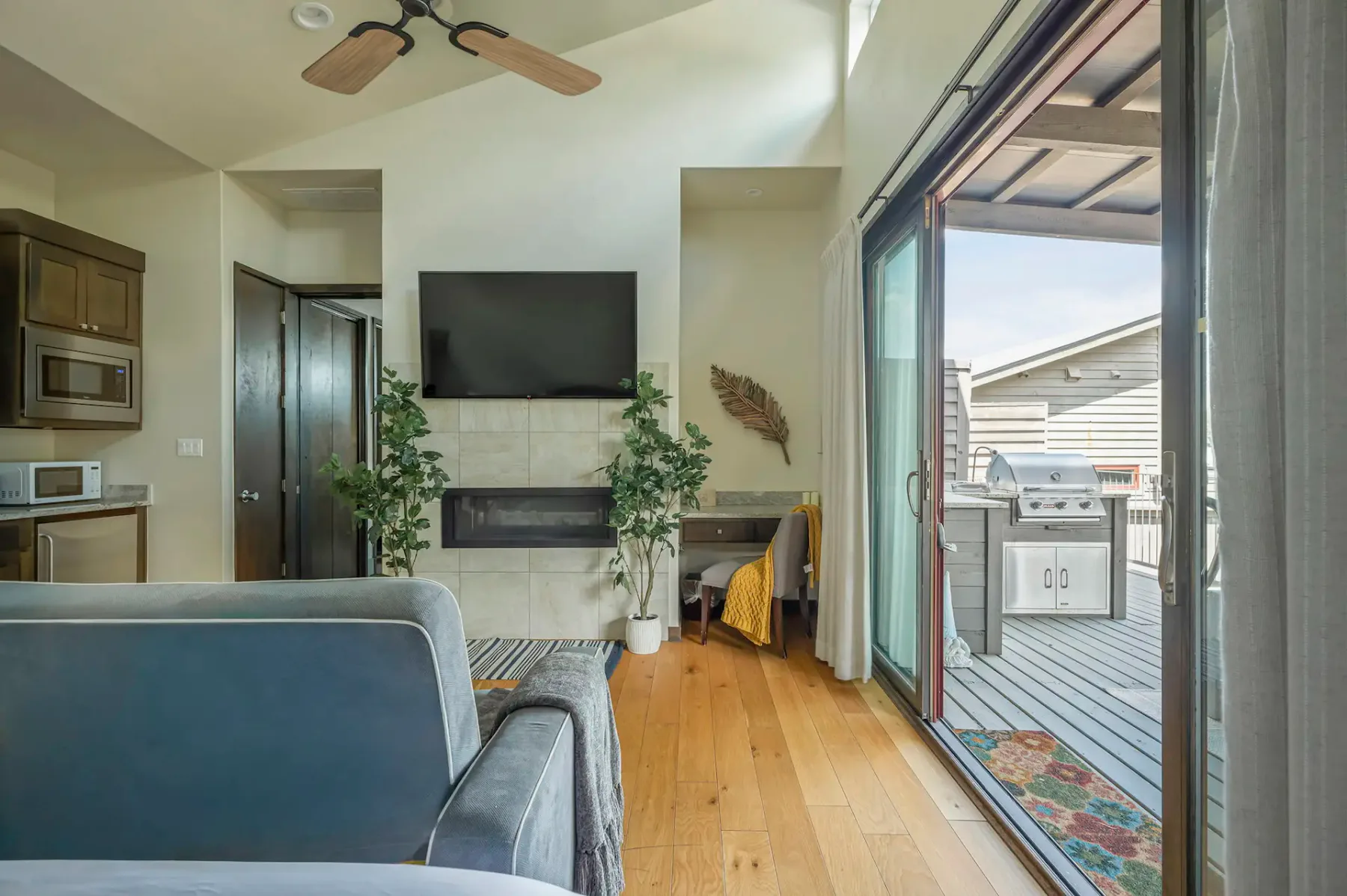 A bright living room with a ceiling fan, wall-mounted TV above a fireplace, plants, and an open sliding door leading to a deck with a grill.