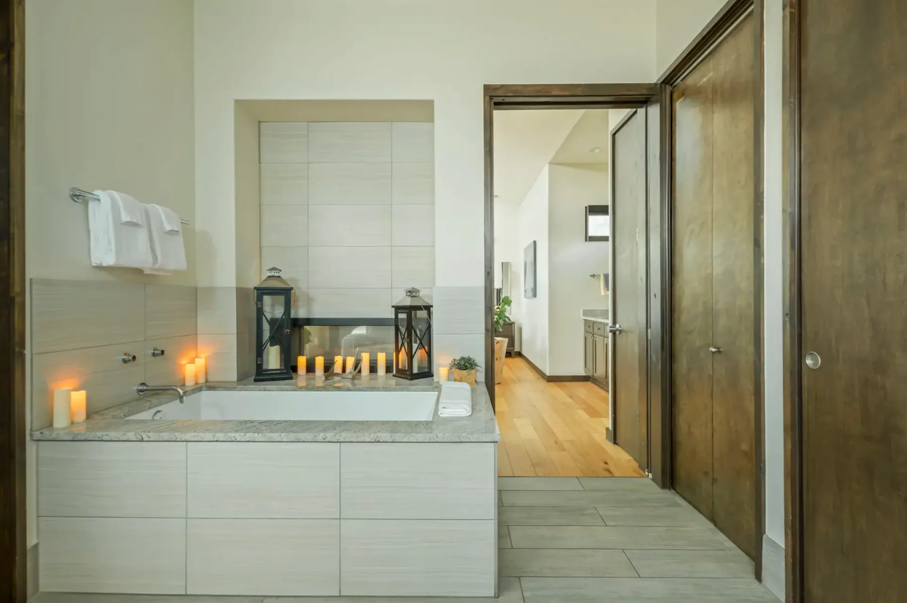 A bathroom with a bathtub surrounded by candles and lanterns, wooden doors, a towel rack, and access to an adjoining room with wooden floors.