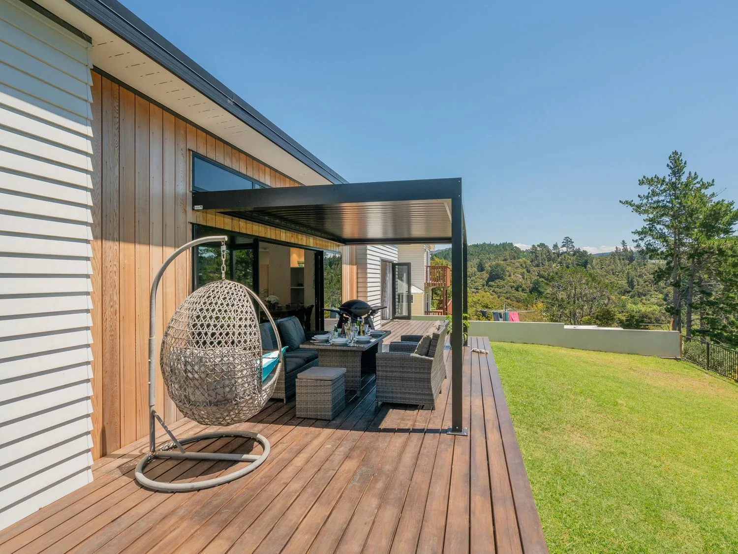 Una terraza exterior de madera con una zona de estar con silla colgante, mesa de comedor y parrilla, con vistas al césped y los árboles en Treetop Bach - Whangamata Holiday Home, Whangamata