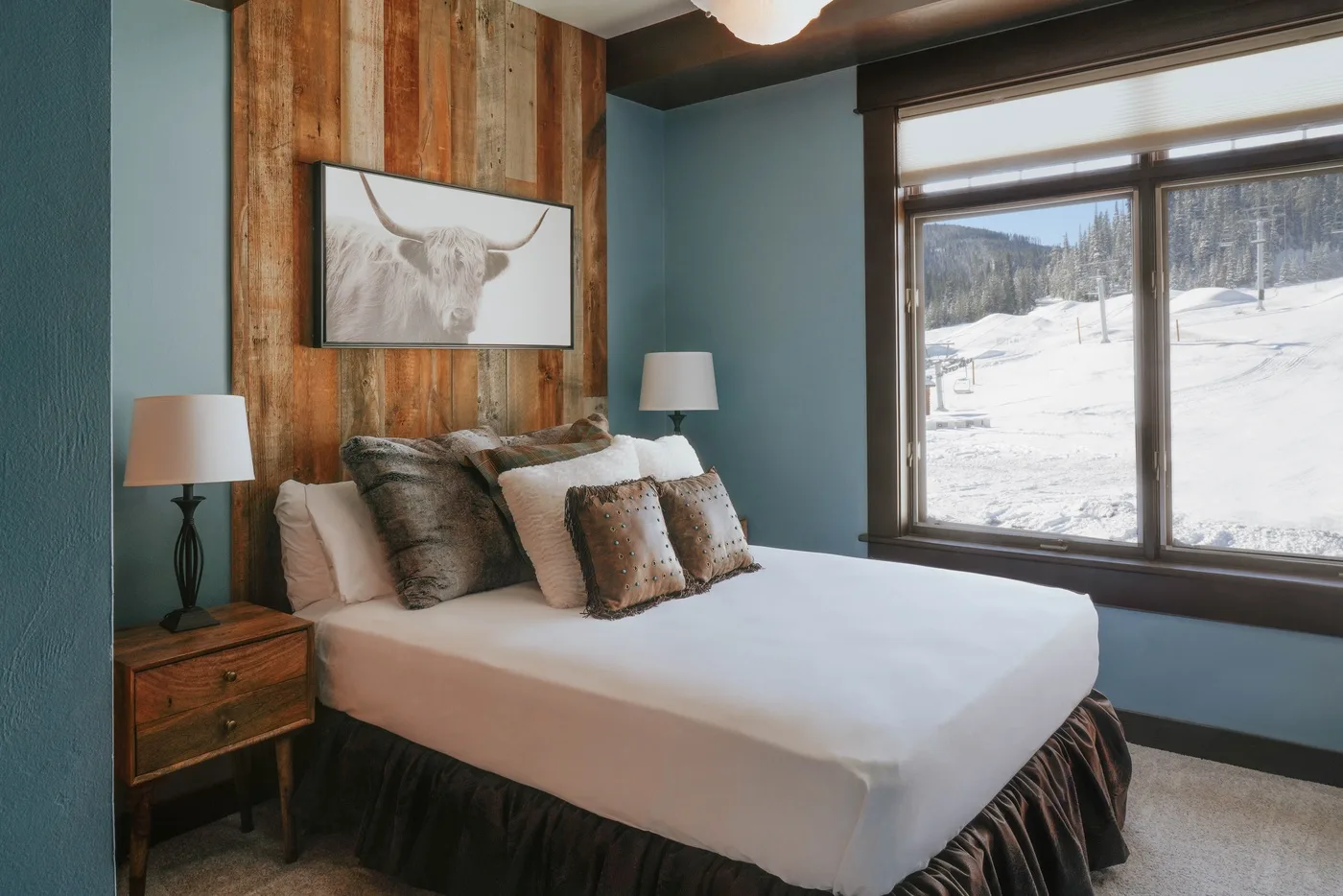A bedroom with a neatly made bed and a large window overlooking a snowy landscape.
