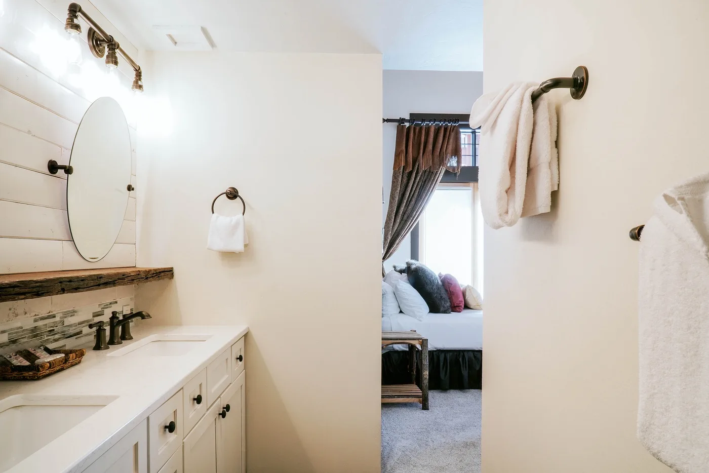 A bathroom vanity with a mirror opens toward a bedroom with a bed.